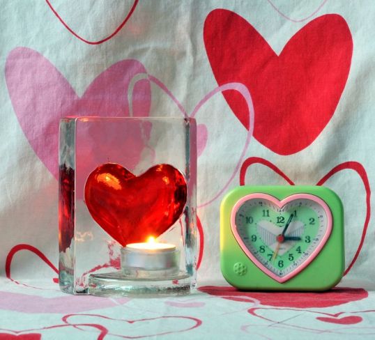 Kynttilälyhty, jossa punainen sydän, ja herätyskello, jossa sydämen muotoinen kellotaulu. Taustalla sydämillä kuvioitu pussilakana.