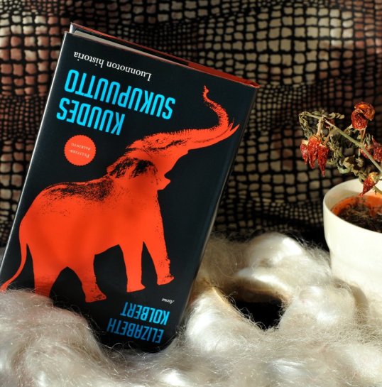 Vasemmalla Kuudes sukupuutto -kirja, jonka kannessa on norsu. Oikealla kuihtunut chilikasvi. Välissä tekoparran suuaukko.