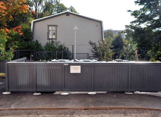 Harmaa aitaus, jonka takaa pilkistää harmaita jäteastioita.