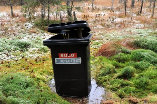 Suon keskellä roskapönttö, johon kiinnitetyt kolme palavaa kynttilää ovat juuri sammumassa pöntön sisältä tapahtuvan puhalluksen voimasta.