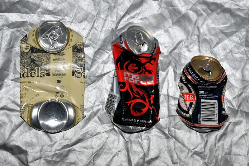 Hopeanvärisen peitteen päällä kolme juomatölkkiä, joissa ylä- ja alaosa ovat kääntyneet pystyyn. Vasemmanpuoleisessa molemmat osat näkyvissä, kahdessa muussa yläosa näkyvissä ja alaosa tölkin vaipan takana.