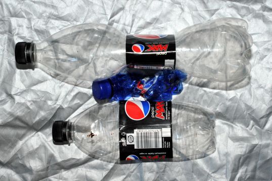 Iso ja pieni kokoon painunut väritön muovipullo sekä sininen täysin sykkyrällä oleva muovipullo hopeanvärisen peitteen päällä.