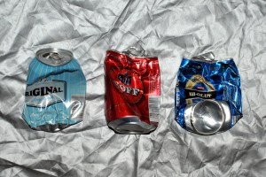 Kolme juomatölkkiä hopeanvärisen peitteen päällä. Vasemmanpuoleinen litistynyt alaosasta, keskimmäinen ja oikeanpuoleinen yläosasta. Oikeanpuoleisessa lisäksi alaosa kääntynyt pystyyn näkyviin.