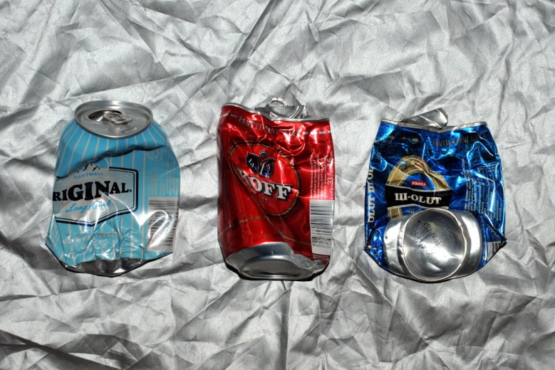 Kolme juomatölkkiä hopeanvärisen peitteen päällä. Vasemmanpuoleinen litistynyt alaosasta, keskimmäinen ja oikeanpuoleinen yläosasta. Oikeanpuoleisessa lisäksi alaosa kääntynyt pystyyn näkyviin.