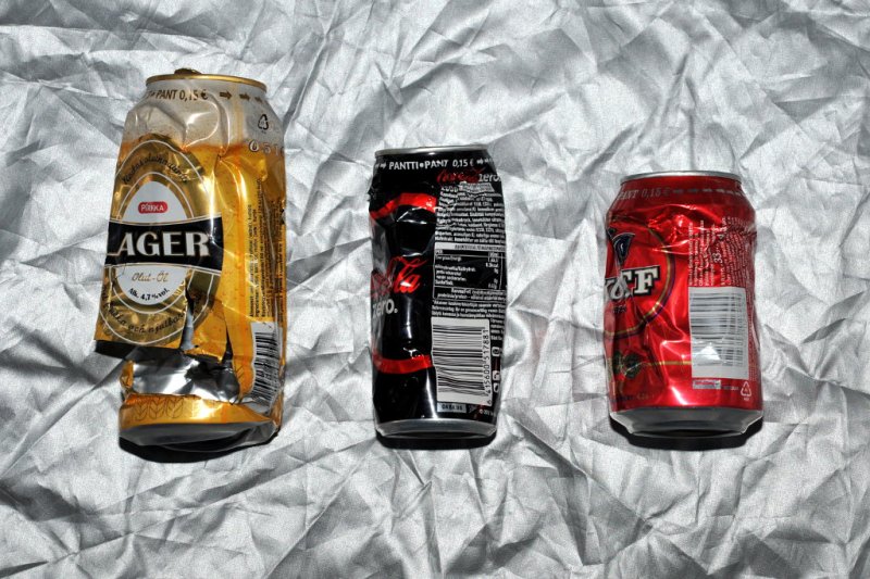 Kolme lommoista juomatölkkiä hopeanvärisen peitteen päällä. Vasemmanpuoleissa tölkissä alaosaa leikattu auki.