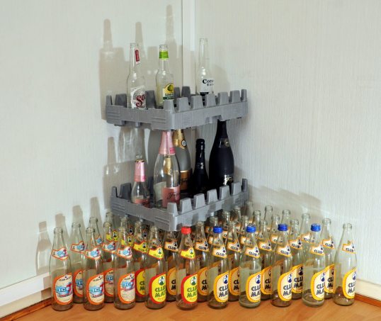 Lattialla Club-Mate-virvoitusjuomapulloja, niiden päällä kennolevy, sen päällä kolme isoa ja kolme pientä Freixenet-kuohuviinipulloa, niiden päällä kennolevy, sen päällä Sol-olutpullo, Somersby-siideripullo ja Corona-olutpullo.