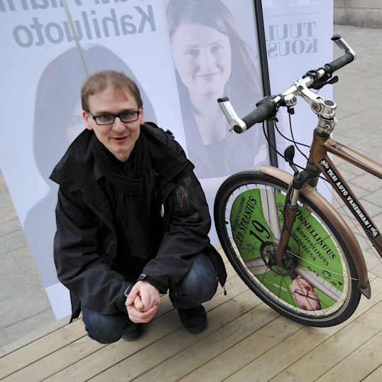 Leo Stranius oman vaalipyöränsä vieressä ja Outi Alanko-Kahiluodon ja Tuuli Kousan vaalijulisteiden edessä.