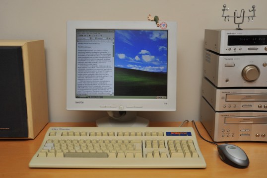 Pöydällä näyttö, stereot, näppäimistö, hiiri ja kaiutin. Näytöllä Windows XP:n työpöytä ja Wordpad-ohjelma, jossa teksti ”Kierrätys on ilomme”. Stereon näytössä näkyy teksti ”Doin’ it Rig”.