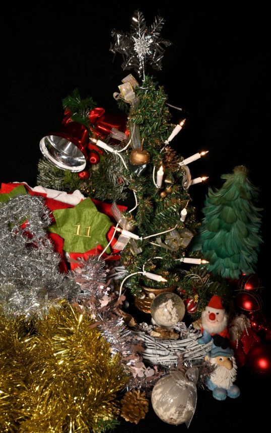 Pieni muovinen joulukuusi, jossa sähkökynttilät. Ympärillä nauhoja, palloja, kelloja ja muita joulukoristeita.